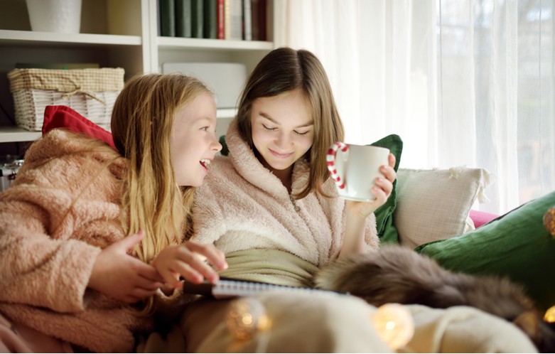 Two people sitting close together on a couch, wearing cozy robes. One person is holding a white mug with a red-striped handle, while the other is pointing at a notebook or tablet resting on their laps. The setting includes soft cushions, a furry blanket or pet, and warm string lights in the foreground, creating a relaxed and festive atmosphere. A bookshelf with books and baskets is visible in the background.