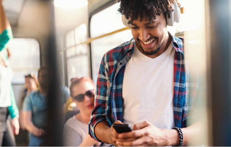 Young man on a bus wearing headphones and smiling at a smart phone.