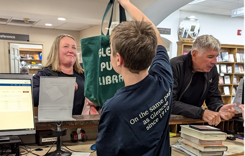 A child at a service desk holds a green tote bag; books and a computer are visible on the counter. The child is wearing a shirt that says "On the same page as Glencoe since 1910".
 
