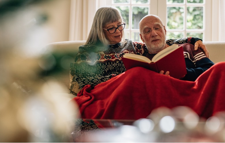 Two people sitting close together on a light-colored couch, wrapped in a bright red blanket. One person is holding an open book, and both are wearing patterned winter sweaters. Behind them is a large window with white framing, showing greenery outside. The foreground is softly blurred, suggesting a cozy indoor setting.