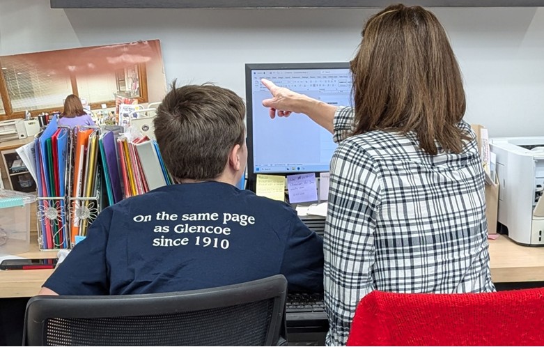 Two people sit at a desk looking at a computer screen; one points at the monitor. A shirt reads “On the same page as Glencoe since 1910.”