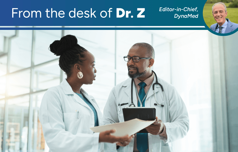 From the desk of Dr. Z. Photo contains two medical professionals wearing white lab coats discussing in the hallway of a hospital. One is holding paperwork while the other was holding a tablet.