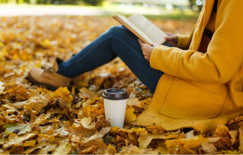 A person in a yellow coat and blue jeans sits on the ground covered in autumn leaves, reading an open book. A disposable coffee cup with a lid rests nearby.
