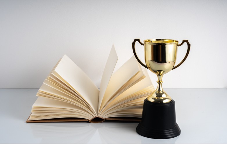 A gold trophy with a black base placed next to an open book with cream-colored pages on a white surface against a plain light background.
