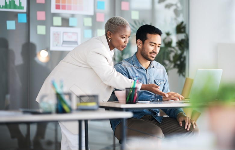 A person in a white suit stands and points at a laptop screen while another person in a denim shirt sits and looks at the screen. They are in a modern office with desks, colorful sticky notes, and charts on a glass wall, suggesting a collaborative work session.