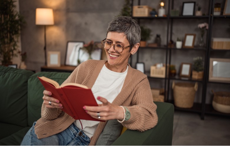 A smiling adult sitting on a green couch reads a red book in a cozy living room. They have short gray hair, wear glasses, and are dressed in a tan cardigan and white shirt. A softly lit lamp and shelves with plants, baskets, and décor are visible in the background.