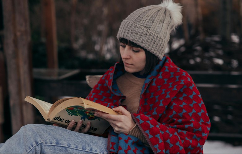 Person sitting outdoors on a bench, wearing a knit hat and wrapped in a red-and-blue patterned blanket, reading an open book.