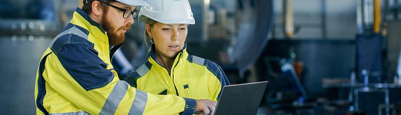 Two workers wearing hard hats and safety jackets sharing a laptop