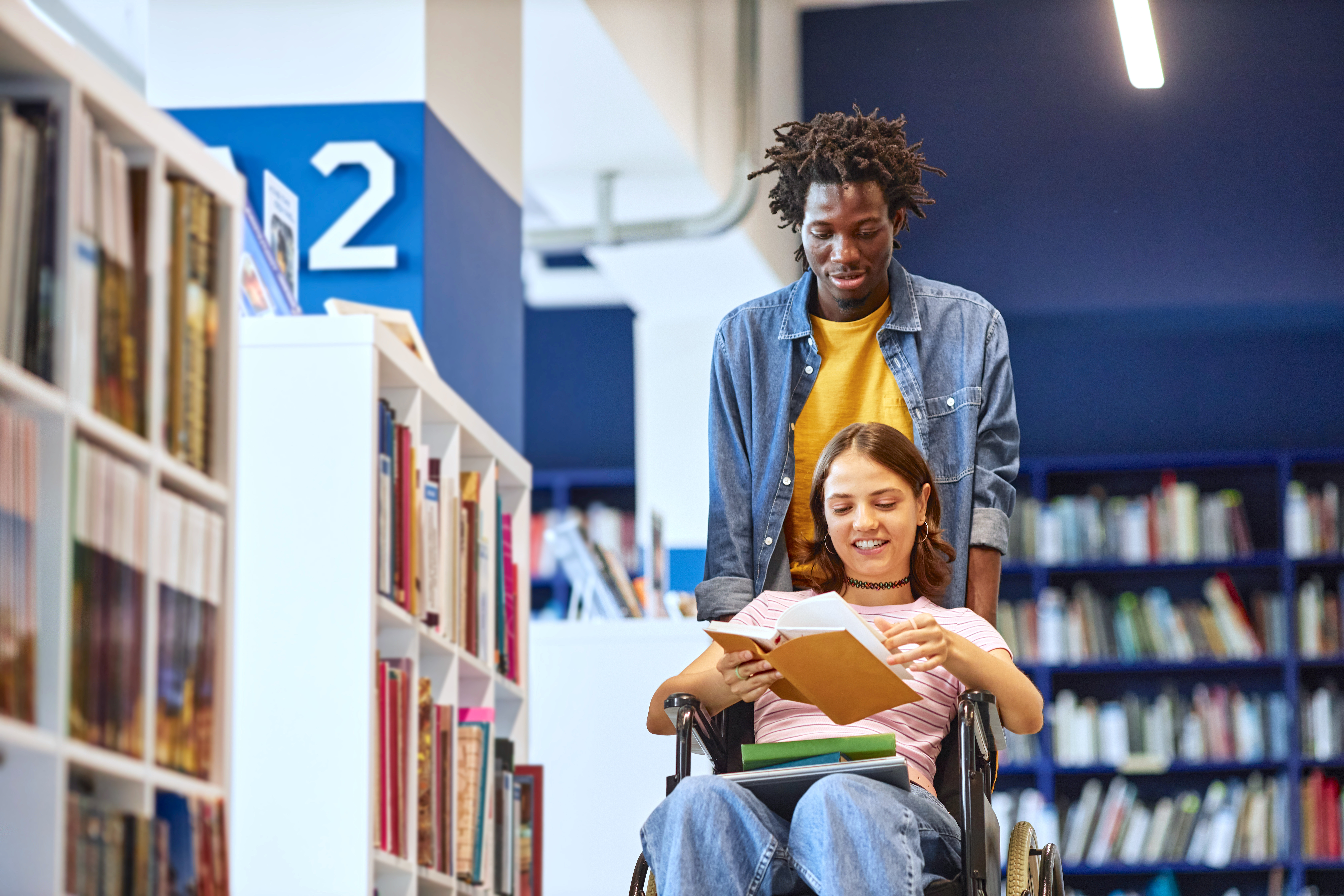 Portrait of young black man assisting female student with disability in college library and pushing wheelchair, copy space