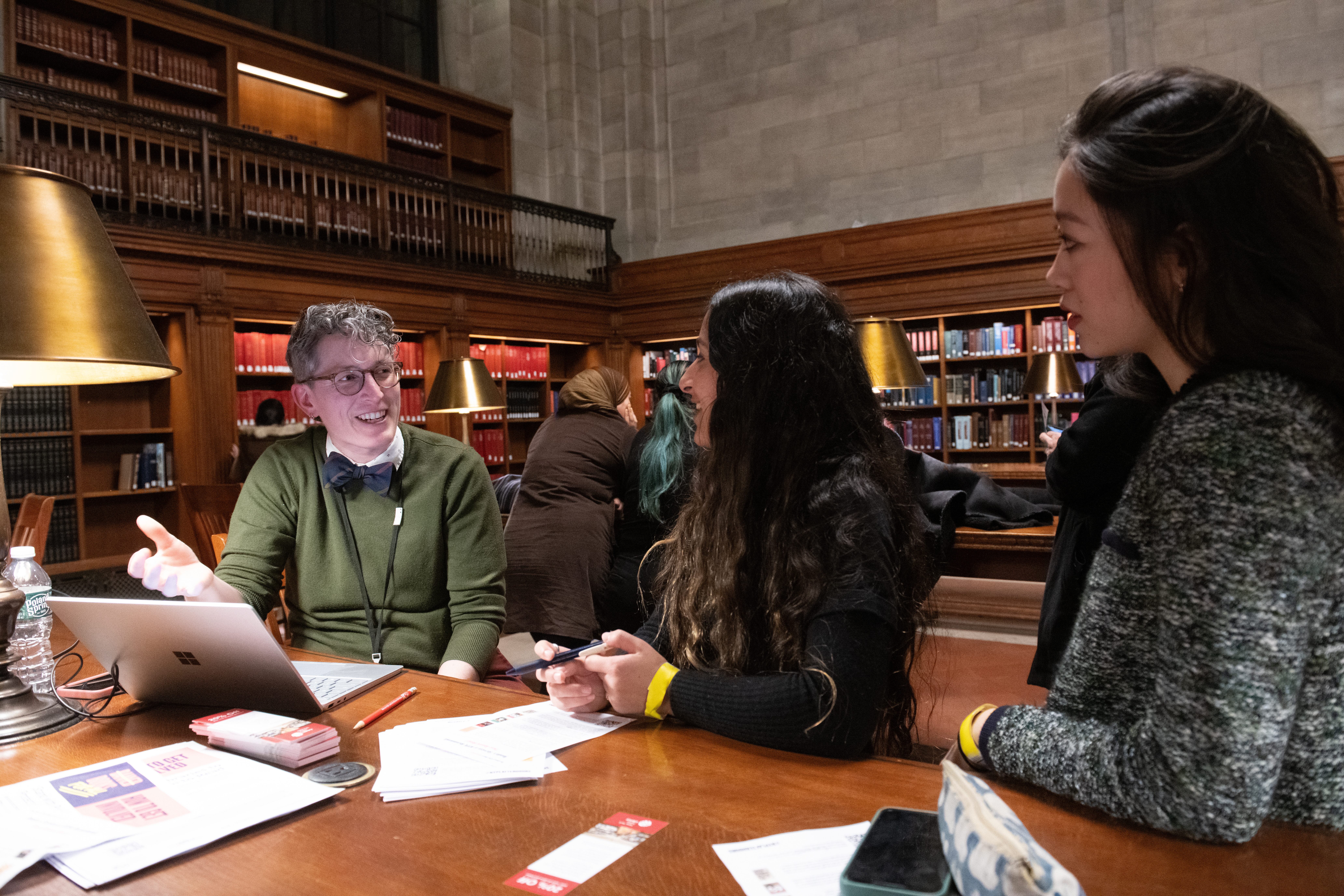 A librarian wearing a green shirt and a bowtie talks to two female patrons in the Rose Reading Room of the New York Public Library