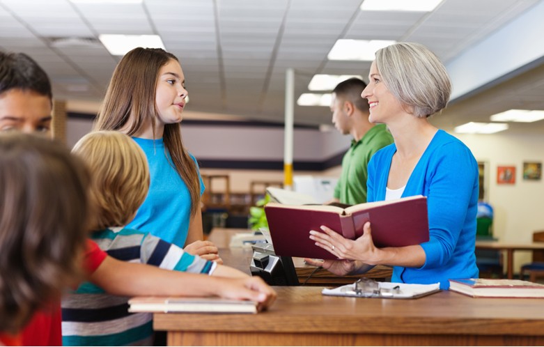 A library setting with a staff member standing behind a wooden service desk, holding an open book and speaking to a group of patrons. The patrons include adults and children gathered around the desk. Books and papers are visible on the counter, and shelves with books are in the background under fluorescent lighting.