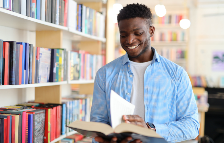 Smiling man reading a book standing next to a library shelf.