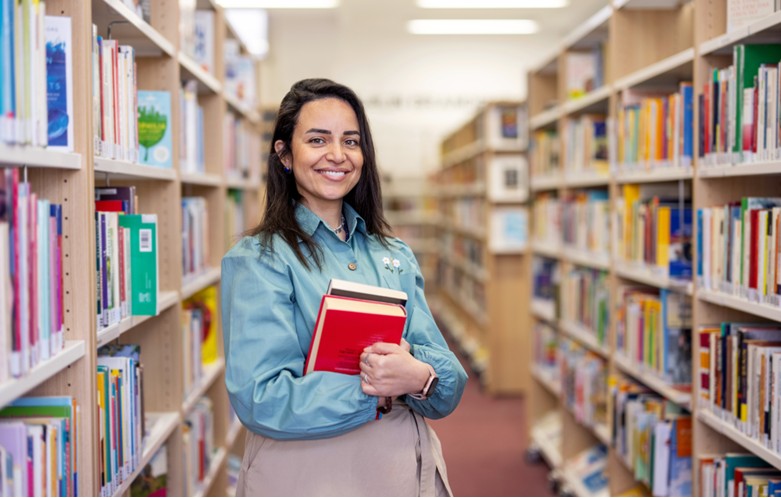 A person standing in a library aisle, smiling at the camera while holding two red-covered books. They are dressed in a light blue shirt and beige pants, surrounded by shelves filled with books.