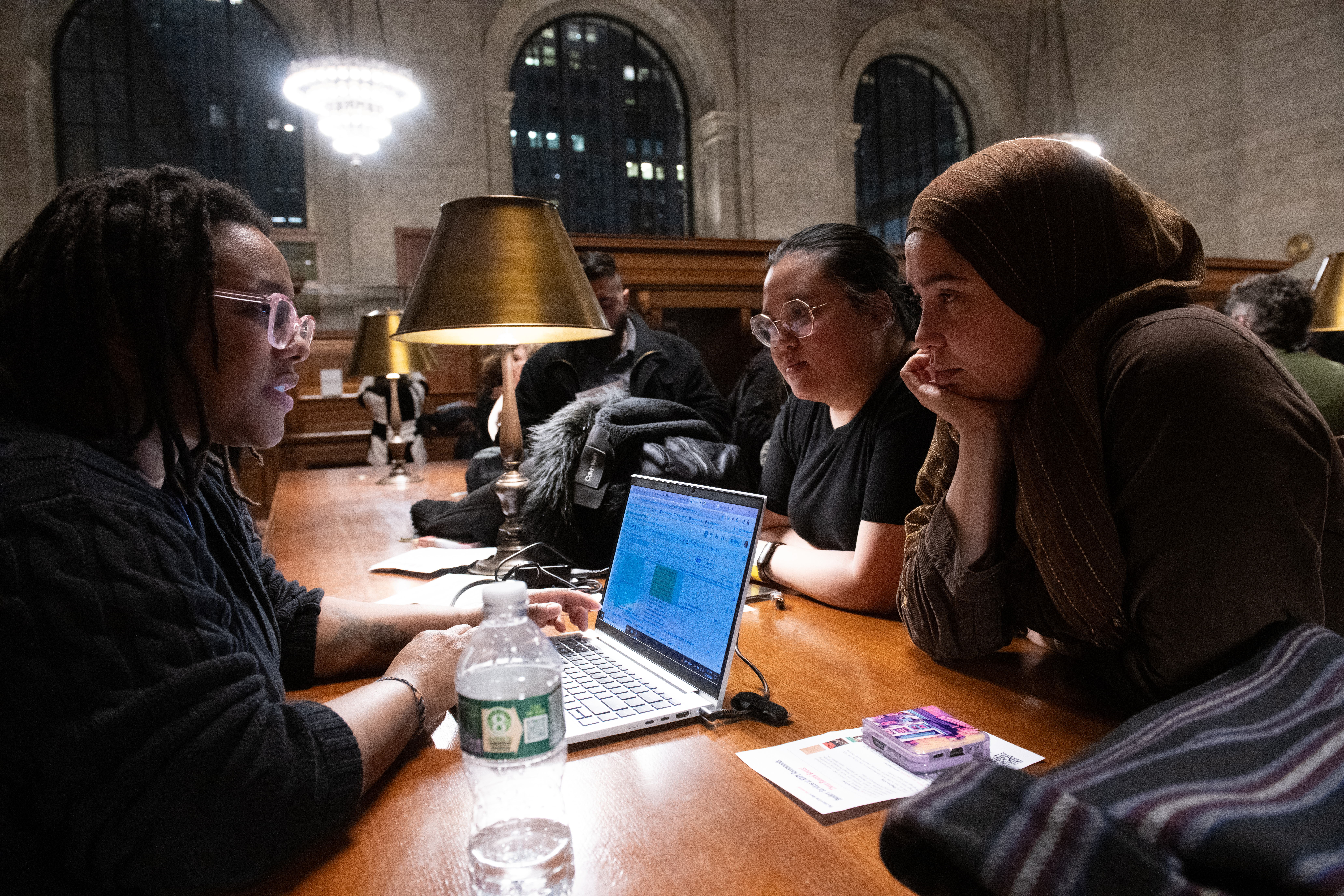 A librarian with a laptop helps to two female patrons in the Rose Reading Room of the New York Public Library