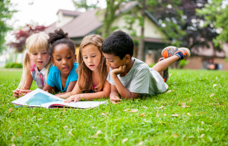 Four children laying in the grass in summer reading a book on a sunny day.