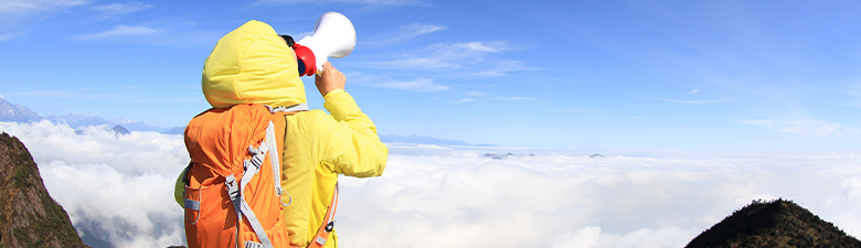 Hiker from behind wearing a hooded jacket and a backpack yelling into a megaphone on a cliff above the clouds