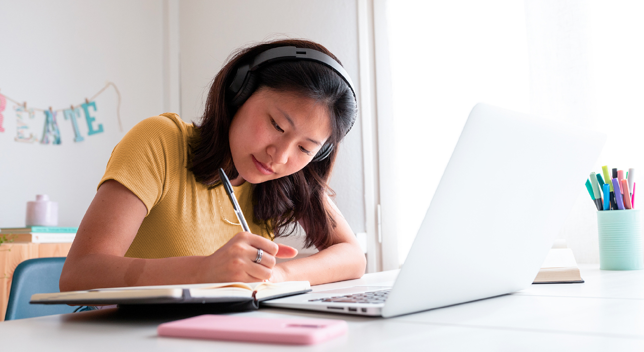 Young student writing in notebook while studying on laptop at desk