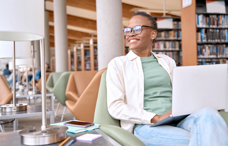 A woman with short hair and glasses wearing a cream-colored shirt layered over a green shirt and jeans reclines in a chair in a library and looks out a window while working on a laptop. 