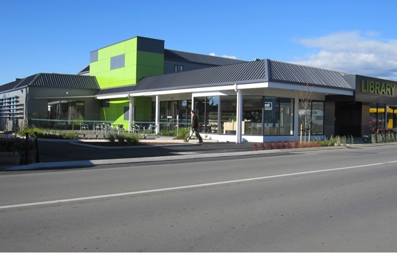 Exterior of a modern public library building with green accents, large windows, and a sidewalk along a street.