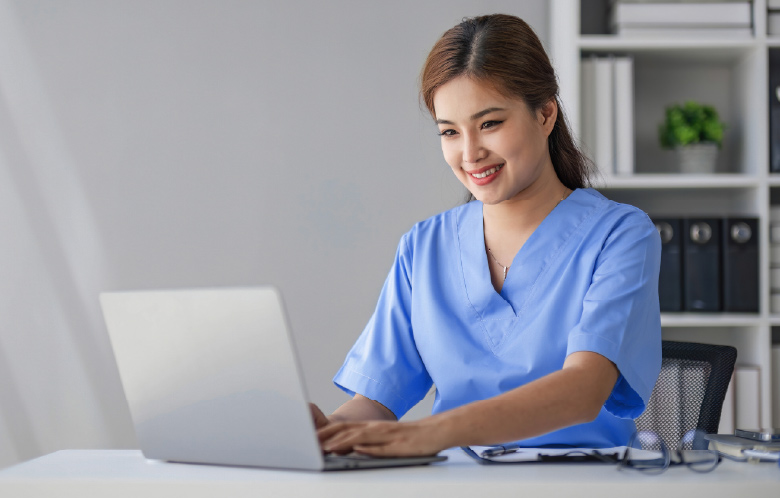 Asian nurse sitting at desk researching on laptop in an office